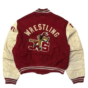 May include: A vintage red and white varsity jacket with a leather sleeve. The jacket has the word "WRESTLING" in a white arc above a cartoon image of an American Indian with the letters "JS" below.