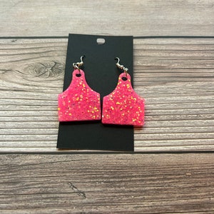 May include: A pair of pink glitter earrings shaped like a cow tag. The earrings are on a black card.