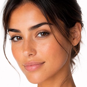 May include: A close-up portrait of a face featuring dark brown eyes, arched eyebrows, and long eyelashes. The skin has a warm, sun-kissed complexion with visible freckles across the nose and cheeks. A small, subtle silver nose stud is visible.