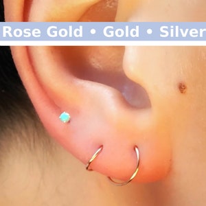 May include: Close-up of a person's ear with three gold hoop earrings and a small turquoise gemstone stud earring. The text "Rose Gold • Gold • Silver" is displayed above the ear.
