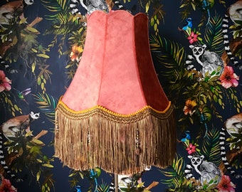 May include: A pink lampshade with a gold fringe trim and a black lamp stand. The lampshade is in front of a dark blue background with a tropical print featuring lemurs, birds, and flowers.