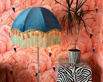 May include: A blue velvet lampshade with a fringe trim sits on a stand in front of a pink flamingo wallpaper.