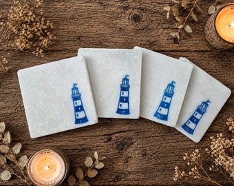 May include: Four square white coasters with a blue lighthouse design. The coasters are arranged on a rustic wooden surface, with a lit candle and dried flowers in the background. The lighthouse design is repeated on each coaster.