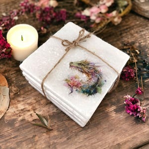 May include: Set of four white square coasters with a watercolor dragon design. The coasters are tied together with twine.