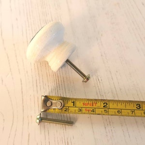 May include: A white cabinet knob with a metal screw attached, positioned next to a yellow measuring tape. The screw measures approximately 1.25 inches in length, while the knob has a diameter of approximately 1.5 inches.