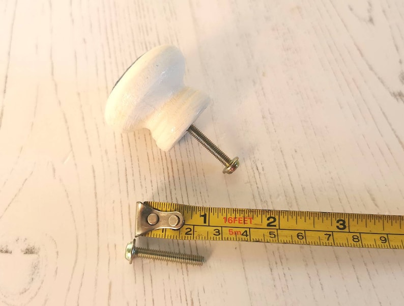 May include: A white cabinet knob with a metal screw attached, positioned next to a yellow measuring tape. The screw measures approximately 1.25 inches in length, while the knob has a diameter of approximately 1.5 inches.