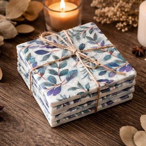 May include: A stack of square coasters with a botanical leaf design in blue and purple, tied with twine. The coasters are on a wooden surface, with candles and decorative elements in the background.