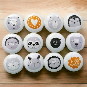 May include: Twelve round white knobs with various animal and word designs. The designs include a sloth, a bear, a penguin, a hedgehog, a heart, a lion, a squirrel, a bunny, a text bubble with "HI" in it, and a speech bubble with "hello world" in it.