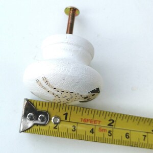 May include: A white wooden cabinet knob with a painted spotted dog design, featuring a metal screw. A yellow measuring tape is positioned along the base, showing the knob has a diameter of approximately 1.5 inches.