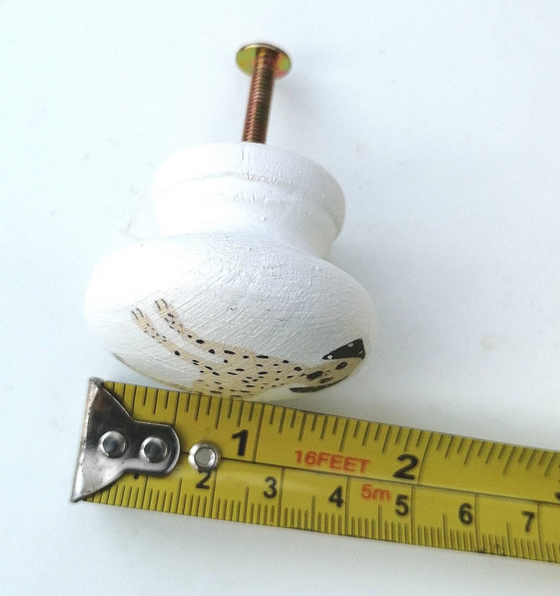 May include: A white wooden cabinet knob with a painted spotted dog design, featuring a metal screw. A yellow measuring tape is positioned along the base, showing the knob has a diameter of approximately 1.5 inches.