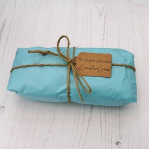 May include: A rectangular package wrapped in light blue paper, tied with brown twine in a bow. A small, rectangular brown paper tag is attached to the twine, featuring the printed text "Handmade with love by CoasterCoast" in black ink.