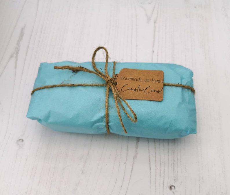 May include: A rectangular package wrapped in light blue paper, tied with brown twine in a bow. A small, rectangular brown paper tag is attached to the twine, featuring the printed text "Handmade with love by CoasterCoast" in black ink.