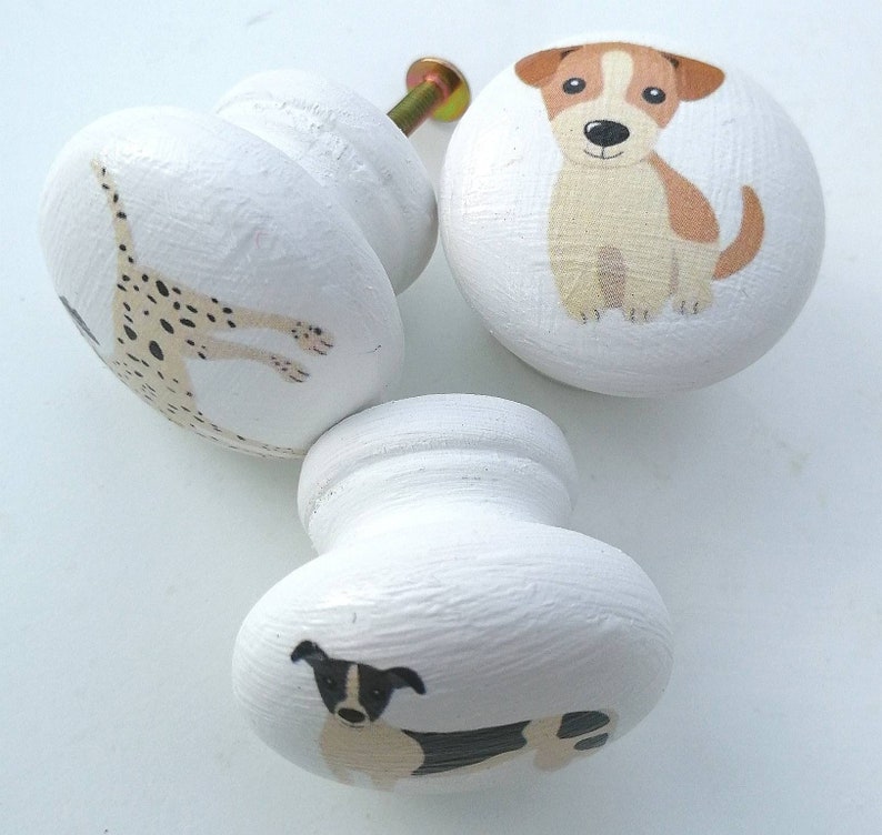 May include: Three round white wooden cabinet knobs, each featuring a different dog illustration. One knob shows a brown and white puppy, another displays a spotted dalmatian, and the third depicts a black and white dog with a metal screw.