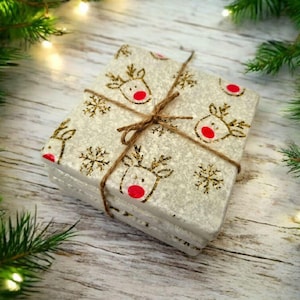 May include: A stack of square coasters tied with twine. The coasters are white with a pattern of gold reindeer and snowflakes with red noses. The background is a wooden surface with Christmas tree branches and lights.