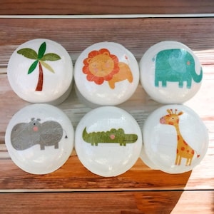 May include: Six white round knobs with different animal and nature designs. The knobs feature a palm tree, a lion, an elephant, a hippopotamus, an alligator, and a giraffe.