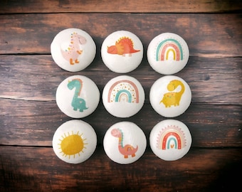 May include: Nine round white painted rocks with different dinosaur and rainbow designs. The designs include a pink dinosaur, a red dinosaur, a green dinosaur, a yellow dinosaur, a rainbow with a heart, a sun, and a rainbow with a heart.