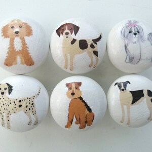 May include: A set of six round white wooden knobs, each featuring a hand-painted illustration of a different dog breed. The dogs depicted include a golden doodle, a brown and white spotted dog, a fluffy gray dog, a dalmatian, a terrier, and a greyhound.