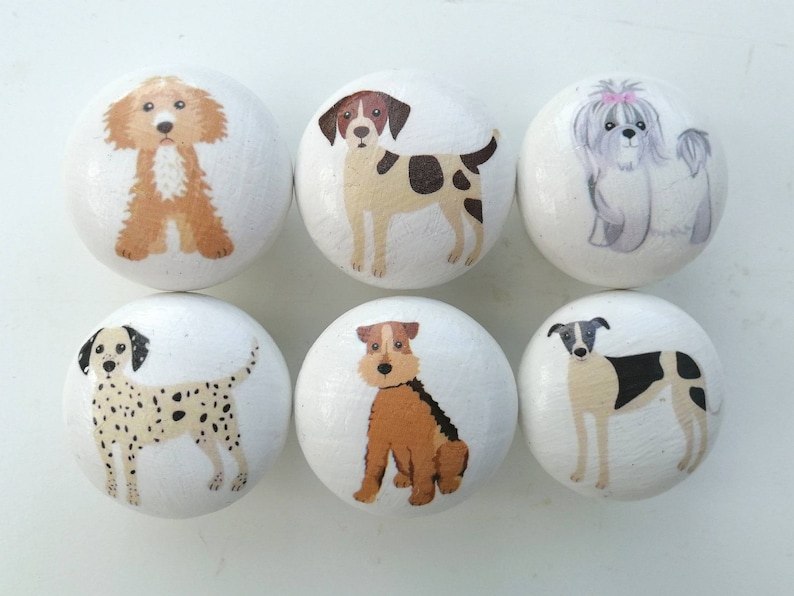 May include: A set of six round white wooden knobs, each featuring a hand-painted illustration of a different dog breed. The dogs depicted include a golden doodle, a brown and white spotted dog, a fluffy gray dog, a dalmatian, a terrier, and a greyhound.