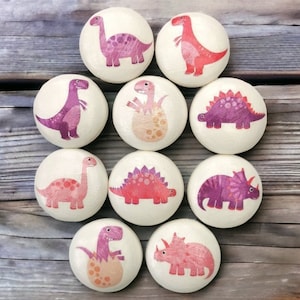 May include: Nine wooden ornaments featuring different dinosaur designs. Each ornament is painted with pink, purple, and red details. The dinosaurs are depicted in various poses, including standing, walking, and hatching from eggs.