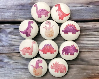 May include: Nine wooden ornaments featuring different dinosaur designs. Each ornament is painted with pink, purple, and red details. The dinosaurs are depicted in various poses, including standing, walking, and hatching from eggs.