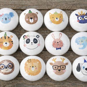 May include: A collection of twelve white ceramic cabinet knobs, each adorned with a different cartoon animal face. The designs include a koala, bear, cat, owl, tiger, panda, rabbit, elephant, sloth, lion, deer, and dog.