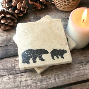 May include: Two beige stone coasters featuring a black bear and cub design. The bear has a moon and stars on its back.