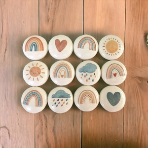 May include: Twelve round wooden ornaments with various designs painted on them. The designs include rainbows, hearts, clouds, suns, and raindrops. The ornaments are all white with a light brown background.