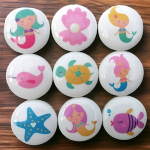 May include: Nine round white ceramic knobs with various sea-themed designs. The designs include a mermaid, a sea turtle, a narwhal, a starfish, a seashell, and a fish.