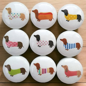 May include: Nine white ceramic knobs featuring dachshund illustrations. Each knob depicts a different dachshund wearing a colourful outfit. The dachshunds are in various poses, including sitting, standing, and lying down.