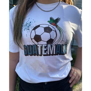 May include: White t-shirt featuring a soccer ball graphic with the word "GUATEMALA" in blue and black. A colorful bird is perched above the ball. The shirt is worn with black pants and a silver chain necklace.