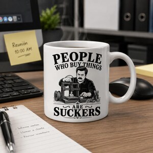 Könnte beinhalten: Ein weißer Keramikbecher mit einer schwarzen grafischen Illustration einer Person, die einen 3D-Drucker bedient. Der Text über der Illustration lautet "PEOPLE WHO BUY THINGS" und der Text darunter "ARE SUCKERS" in fetter, schwarzer Schrift.