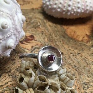 Op de afbeelding: Een zilveren ring met een ronde, platte, zilveren bovenkant en een kleine, donkerrode edelsteen in het midden.