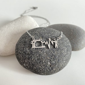 Può includere: Collana d'argento con un ciondolo a forma di cuore che rappresenta una macchina da cucire e delle forbici. La collana è su una roccia grigia.