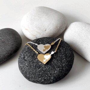 May include: Two silver necklaces with heart-shaped pendants. The pendants are interlocking and feature an arrow piercing through the centre. The necklaces are displayed on a grey stone.