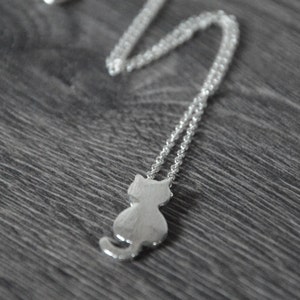 May include: A silver chain necklace with a silver cat pendant. The cat is facing away from the viewer and has a simple, minimalist design.