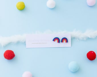 Op de afbeelding: Regenboogvormige oorbellen met turquoise, oranje en paarse accenten, gepresenteerd op een witte kaart. De kaart ligt op een lichtblauwe ondergrond, omringd door kleurrijke pompons en een witte verenboa. Een kleurrijk accessoire.