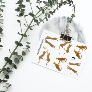 Puede incluir: Una hoja de pegatinas de l&aacute;mina dorada con un patr&oacute;n repetitivo de tigres estilizados en varias poses. Las pegatinas est&aacute;n sobre un fondo blanco y tienen el texto "eyesdrop designs." impreso en negro en la parte superior.