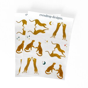 Puede incluir: Una hoja de pegatinas con varias poses de leopardos en color dorado. Las pegatinas est&aacute;n sobre un fondo blanco con algunas estrellas y una luna creciente. El texto "eyesdrop designs." est&aacute; en la parte superior de la hoja.