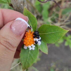 以下が含まれることがあります： オレンジ、黒、白のビーズで作られた蜂のチャーム。緑の葉を背景に持っています。チャームは小さなビーズで作られており、銀色の留め金が付いています。蜂の体はオレンジ色で、黒と白のアクセントがあります。