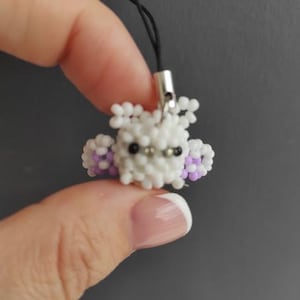 May include: A handmade beaded charm in the shape of a small, white owl with black eyes and purple accents. The owl charm is attached to a black cord with a silver-colored clasp.