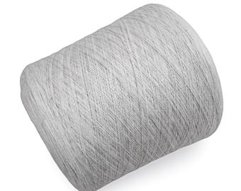 Puede incluir: Una gran bobina de hilo gris claro, bien enrollada y ligeramente inclinada hacia la izquierda. El hilo parece suave y fino, adecuado para proyectos de tejido o ganchillo. El fondo es blanco.