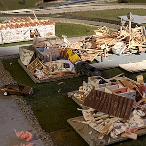 May include: A model of a house destroyed by a natural disaster. The house is made of cardboard and is in pieces. There are also small toy cars in the image.
