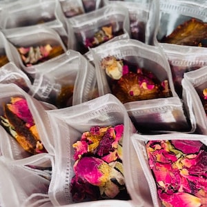 Könnte beinhalten: Nahaufnahme von mehreren kleinen weißen Netzsäckchen, gefüllt mit getrockneten Rosenblüten und anderen Pflanzen. Die Säckchen sind in einem Gittermuster angeordnet und bilden so eine optisch ansprechende Darstellung.