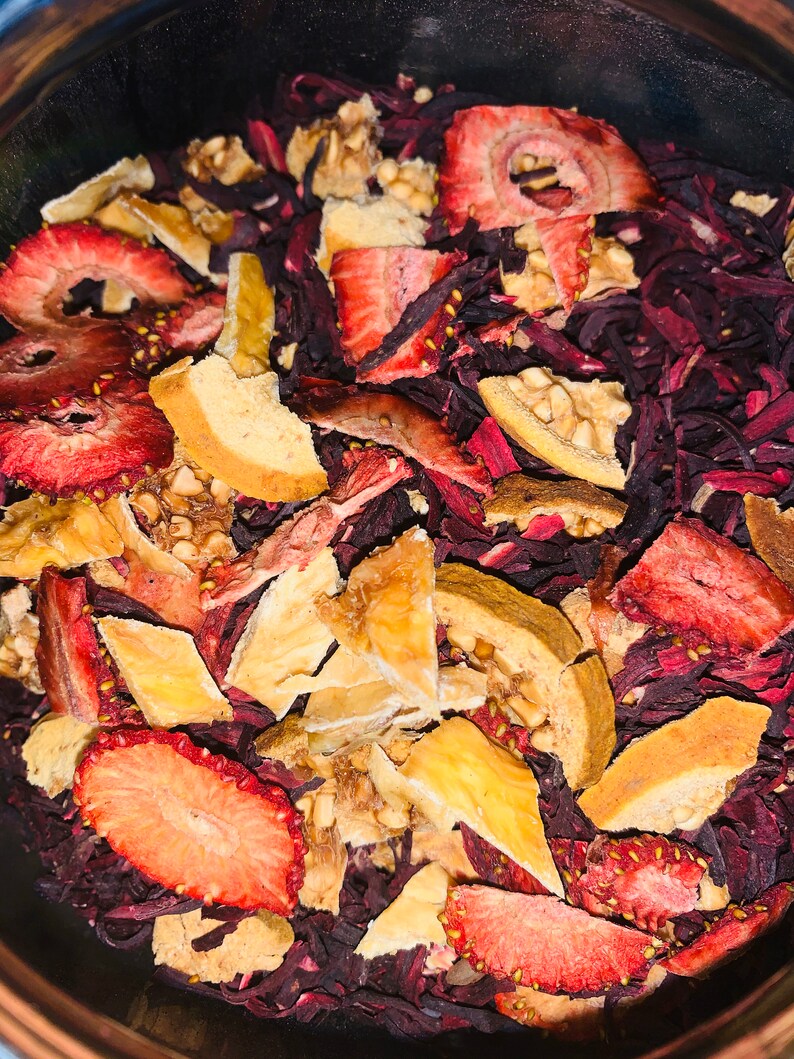 May include: A close-up of a bowl filled with dried fruit and flower petals. The fruit is a mix of red strawberries and light brown pieces of dried fruit. The flower petals are a deep red color.