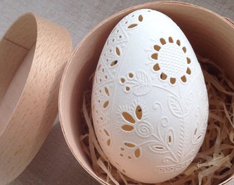 May include: A white, intricately carved egg with a floral design sits inside a round wooden box lined with shredded paper.