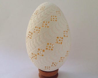 May include: A decorative, off-white egg with intricate, carved leaf and square patterns. The egg sits on a small, round, wooden stand. The egg's interior is a warm yellow color, visible through the cut-out designs.