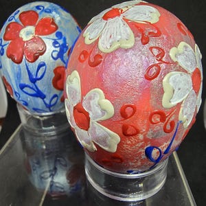 May include: Two decorative egg-shaped ornaments featuring raised paint designs. One egg has a light blue base with red flowers and blue swirls. The other egg has a red base with white flowers and red accents. Both are displayed on clear stands.