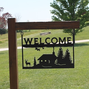 May include: A black metal welcome sign with a cabin, trees, and a deer silhouette. The sign is hanging from a wooden post.
