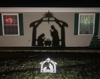 May include: A white nativity scene silhouette with a star on top, illuminated by a spotlight, is displayed on a lawn in front of a house with Christmas lights in the windows.