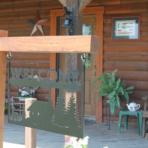 May include: A wooden sign with a metal cutout that says "WELCOME" with a cabin and trees in the background. The sign is hanging from a wooden post in front of a log cabin.